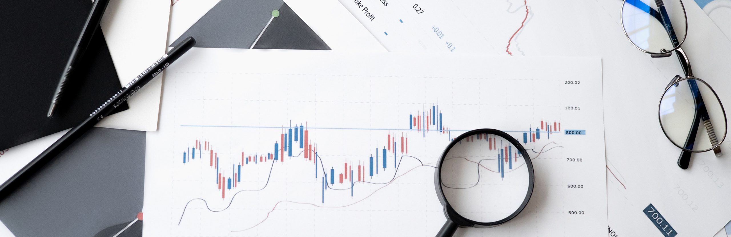 An overhead view of papers with various graphs, a magnifying glass, a pair of glasses, a pencil, and a pen.