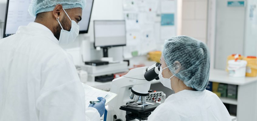 Two people in white protective lab coats, masks, and bouffant caps. One person is standing and holding paperwork. The other person is sitting down and looking through a microscope.