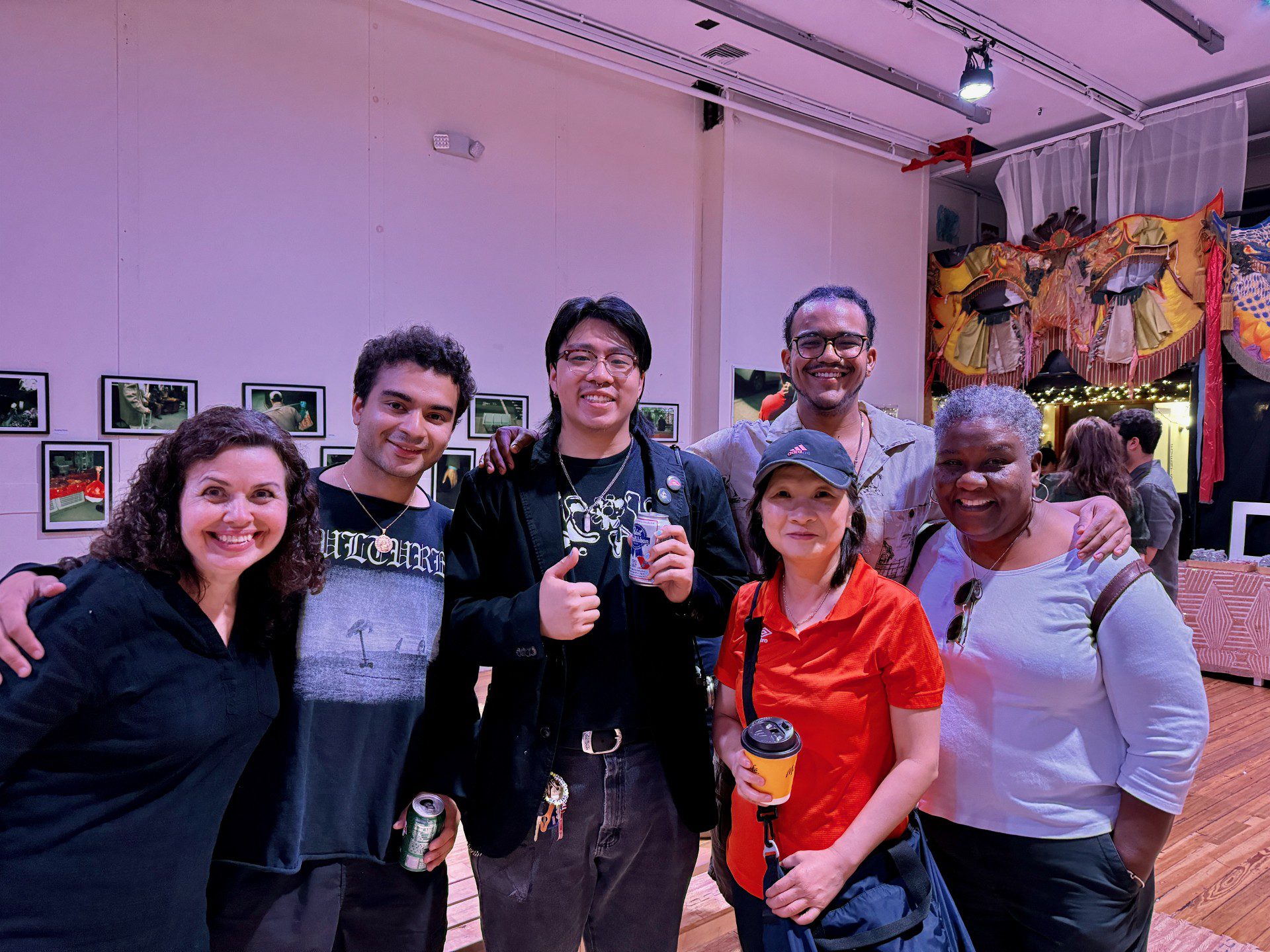 A group of people standing close together, smiling into the camera. They are in a gallery with framed photos behind them.